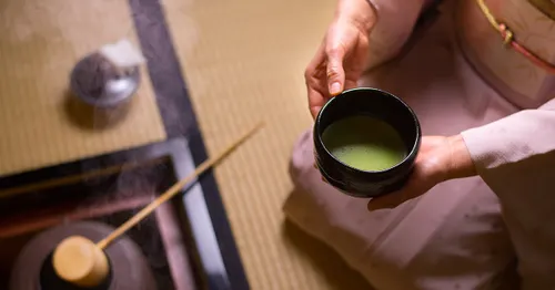 Hands offering a black raku-style bowl of whisked matcha